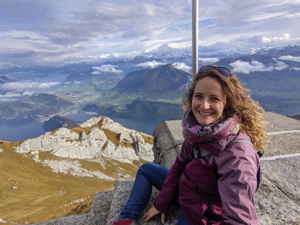 In the foreground a woman sits on mountain overlook in Switzerland with golden fields and rolling green mountains in the background.