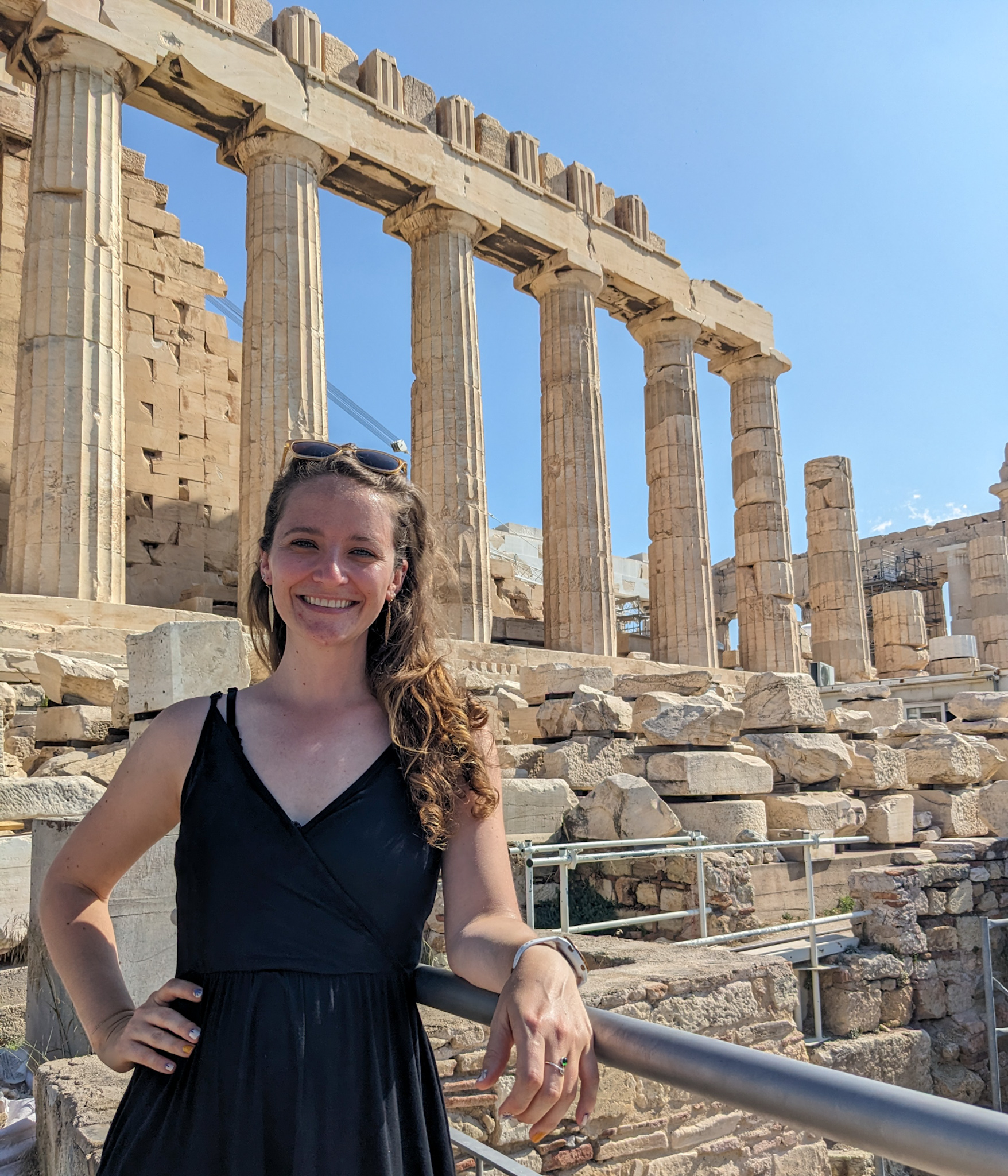 Caucasian woman stands in front of the Parthenon smiling while leaning on a railing with one arm and the other is on her hip