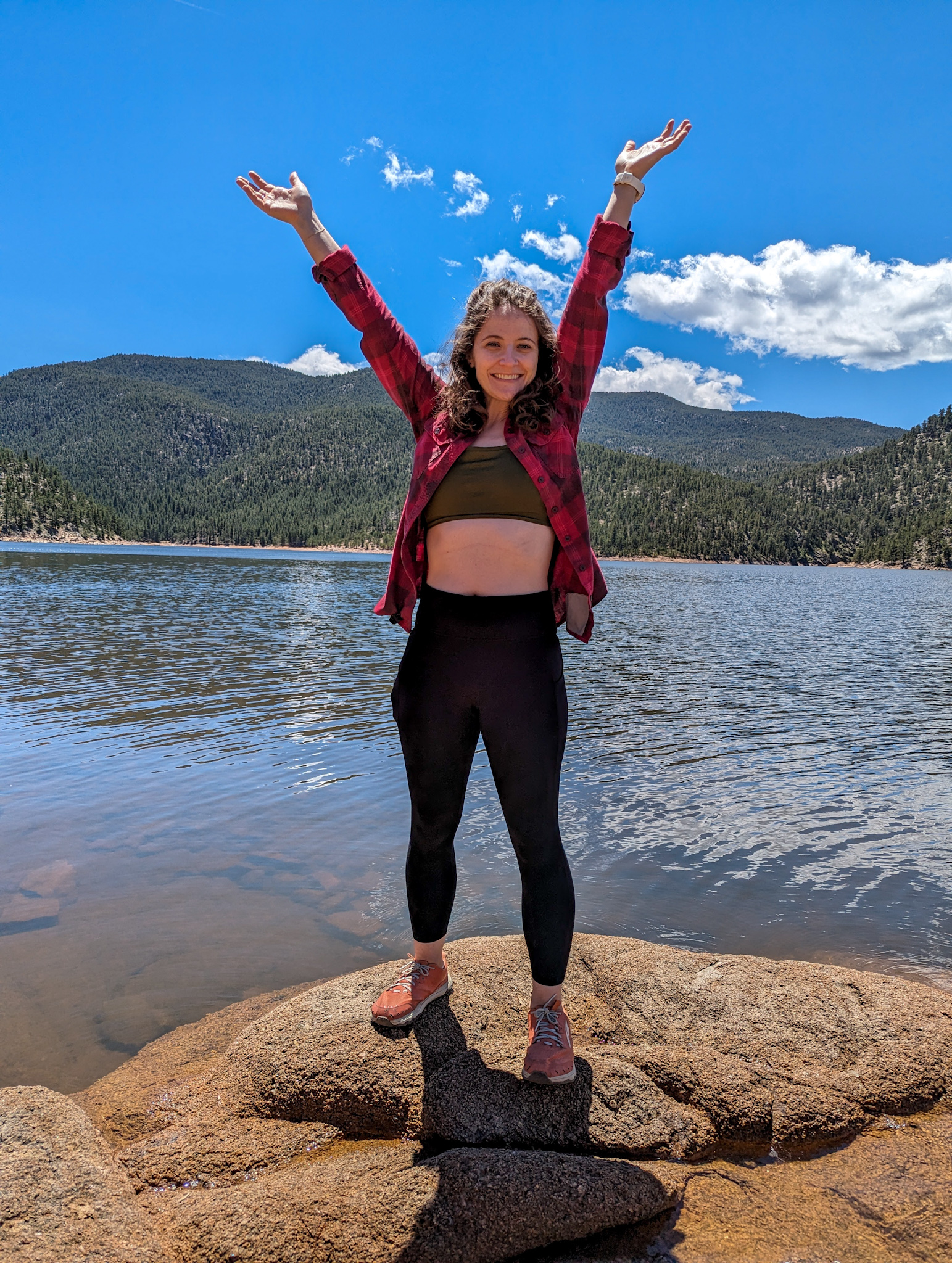 Caucasian women with brown curly hair stand on rock near water with arms in the air smiling